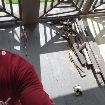 A man in a red shirt is sitting on a porch next to a pile of wood.
