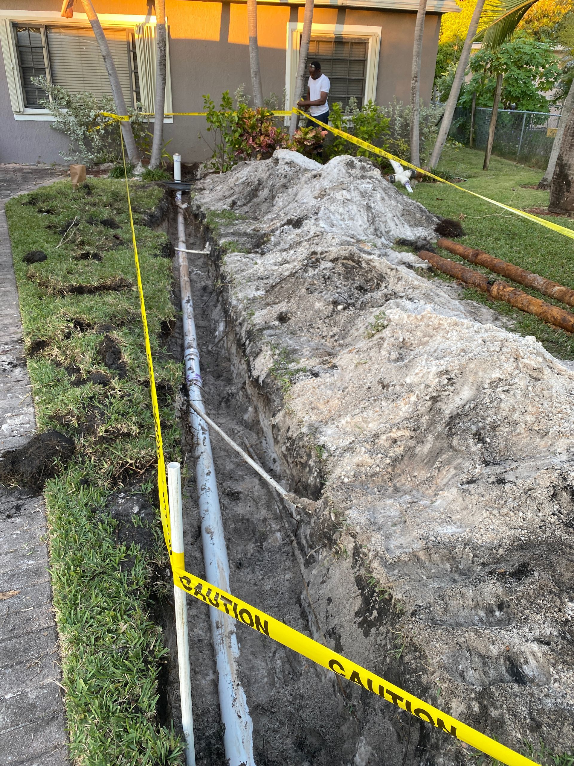 A construction site with a trench, pipes, and caution tape running alongside a house.