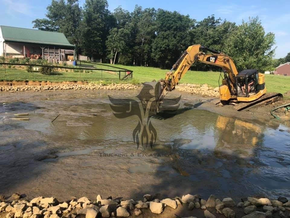 Excavator digging in a muddy pond beside a small cabin and wooded shoreline