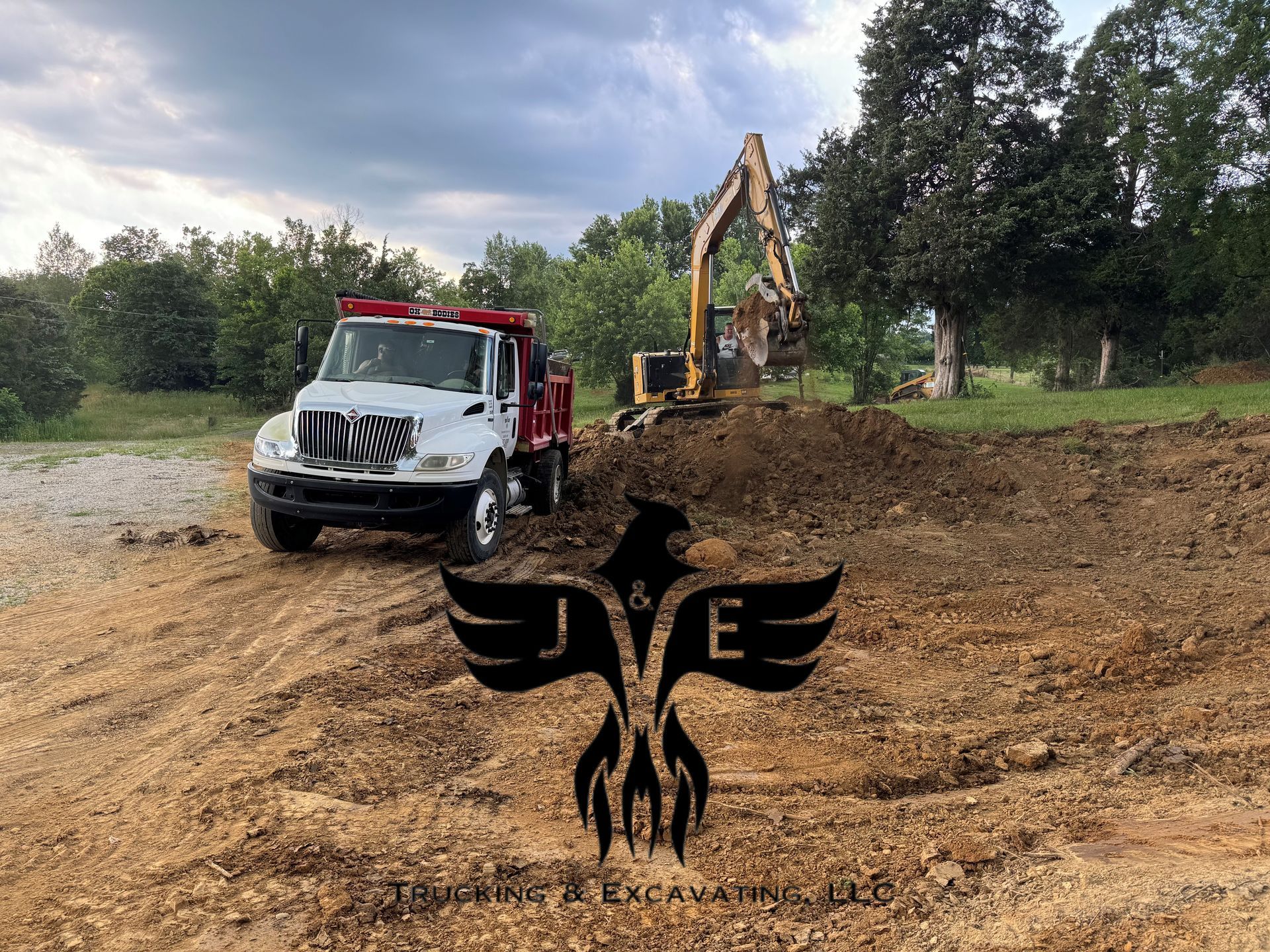 Tow truck and excavator at a muddy roadside worksite with trees and cloudy sky
