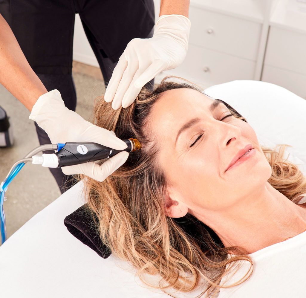 Woman receiving scalp treatment with a device, eyes closed, being assisted by gloved hands.