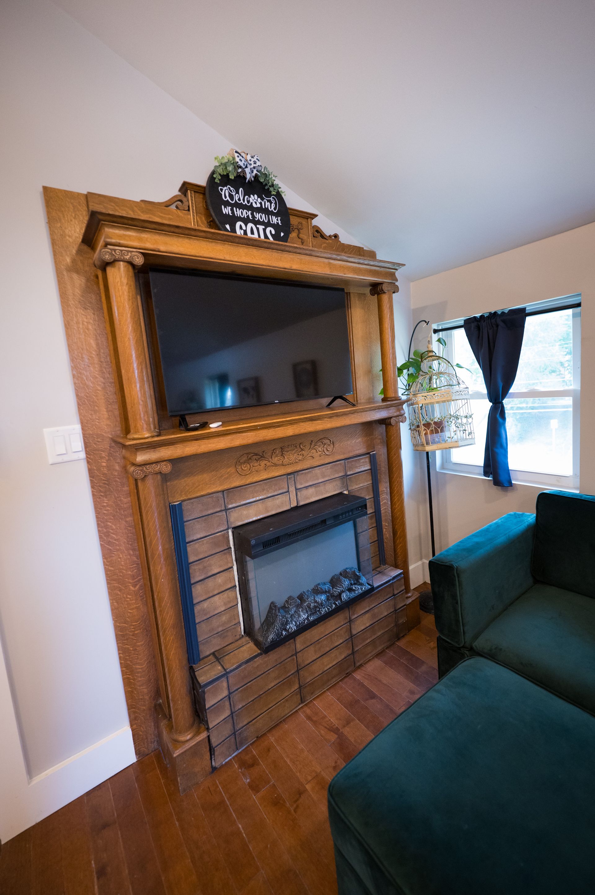 Wooden framed electric fireplace with TV above, green velvet couch, and window.
