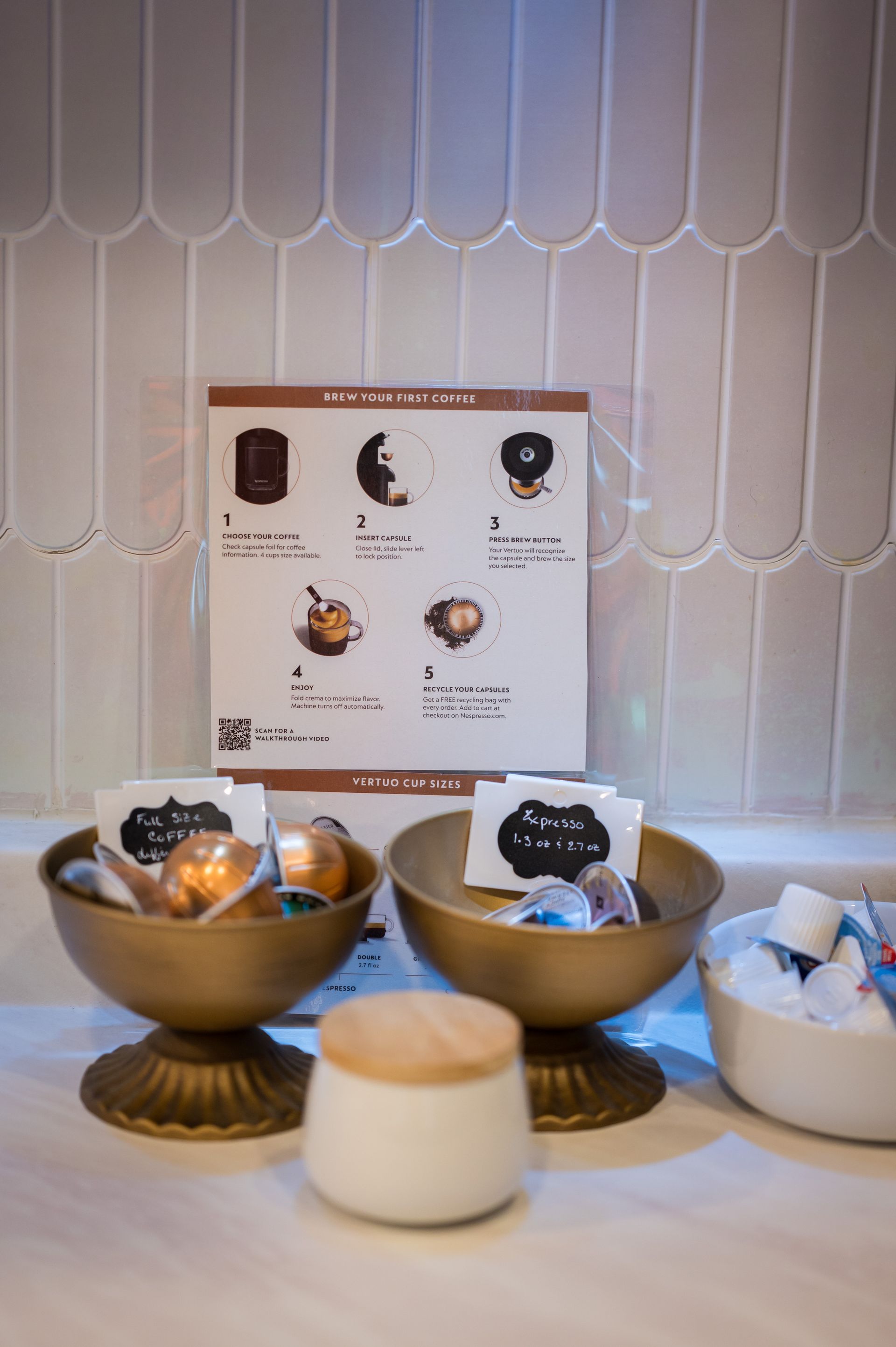 Display of chocolates in golden bowls with a menu sign and small ceramic jar against white tiled wall.