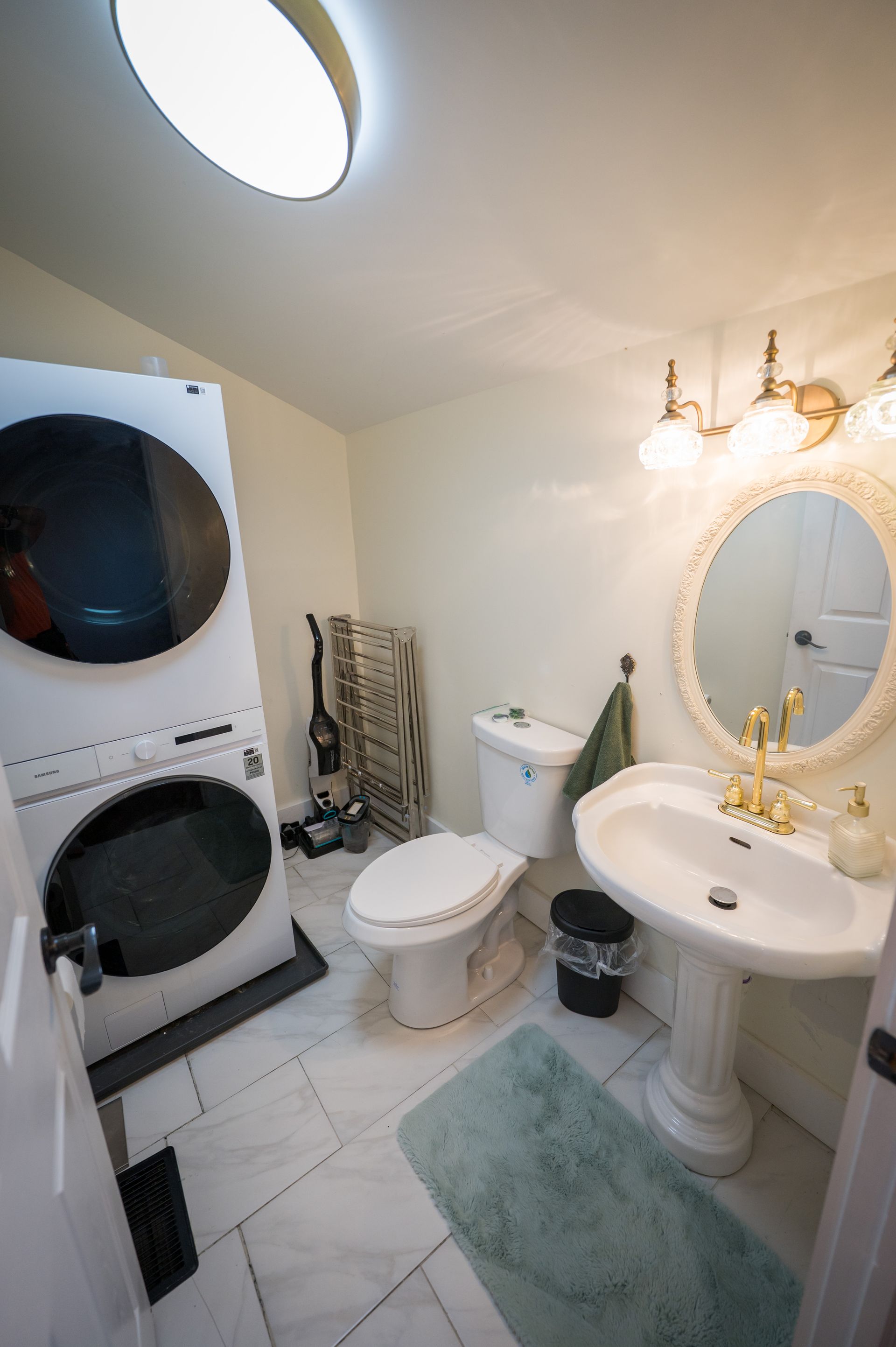 A small bathroom with a stacked washer and dryer, toilet, pedestal sink, and rug.