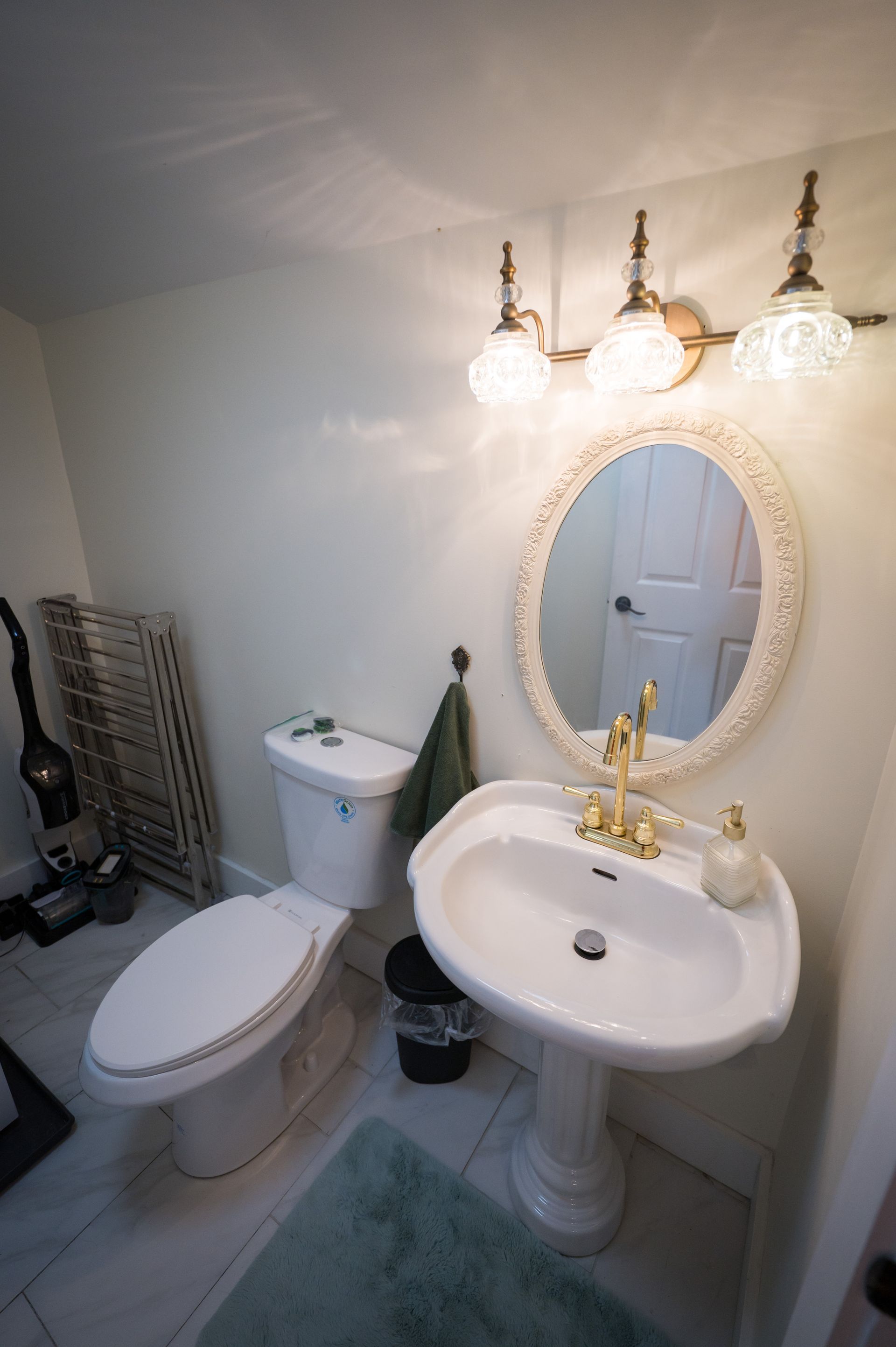 Bathroom with white pedestal sink, toilet, and ornate mirror. Soft green rug and wall color.