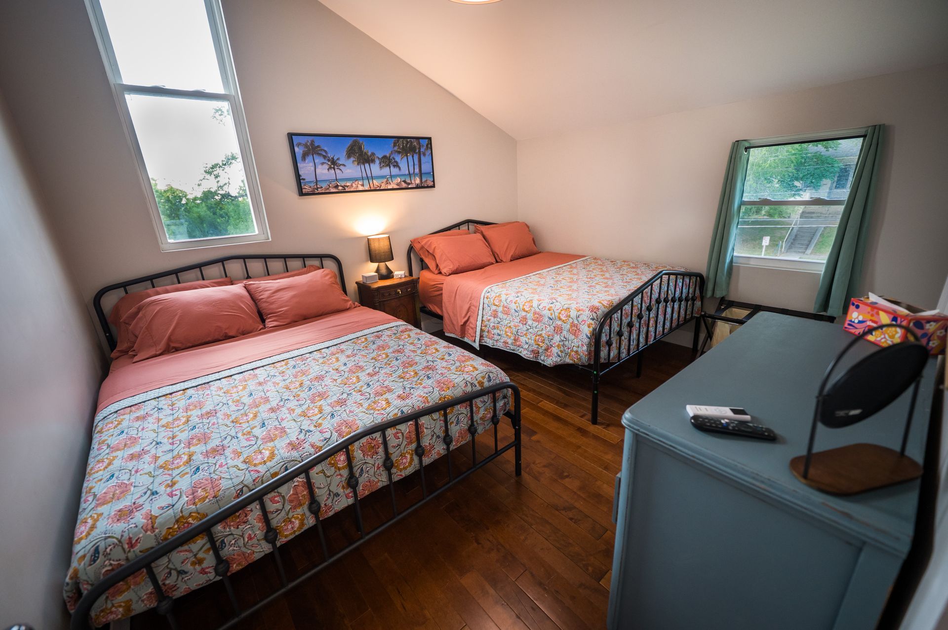 Bedroom with two beds, dresser, window. Peach bedding, dark metal bed frames. Hardwood floors.