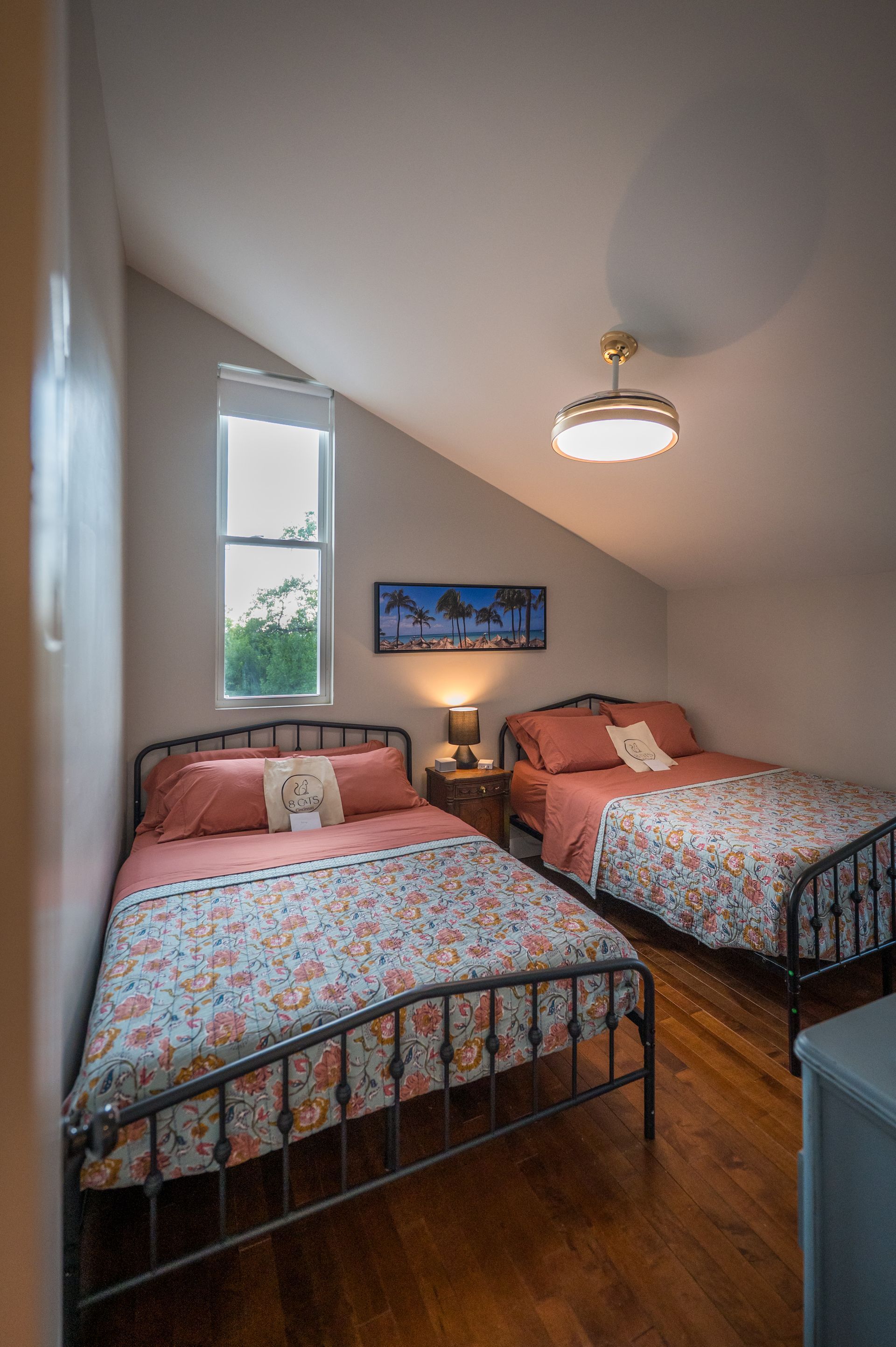 Two beds in a bedroom with slanted ceiling, floral bedspreads, wooden floor, and a small window.