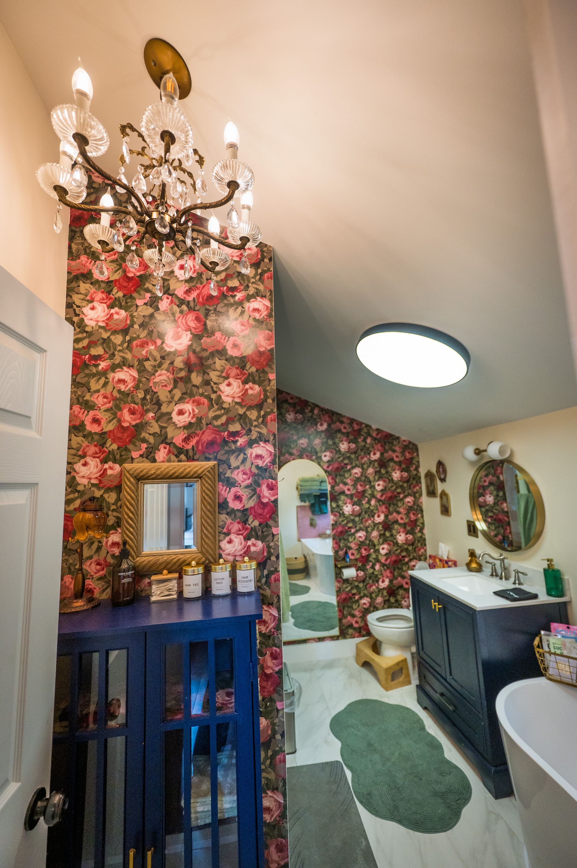 Bathroom with floral wallpaper, chandelier, blue cabinet, and round mirror.
