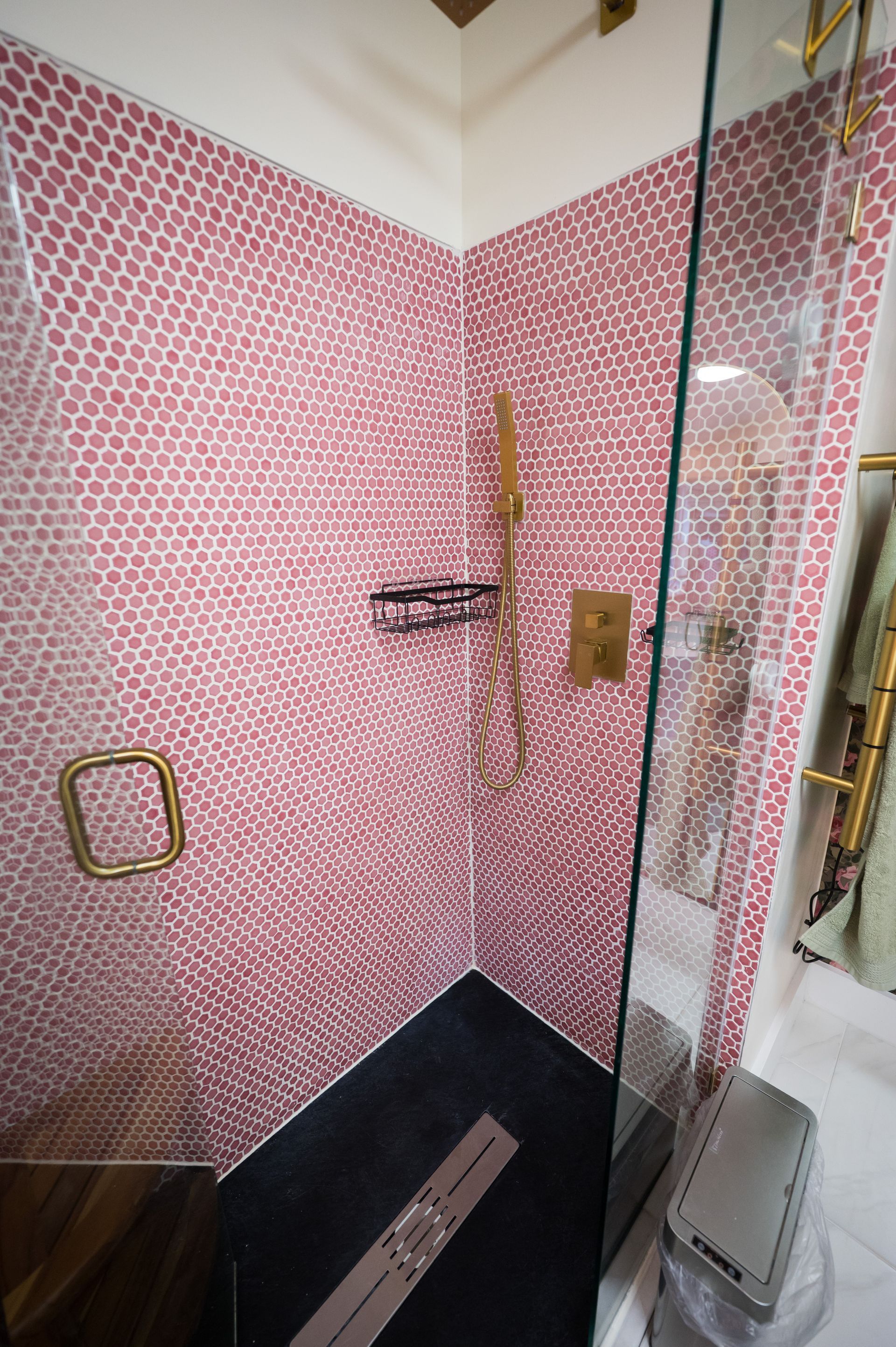 Corner shower with pink penny tile walls, glass door, black floor, and gold fixtures.