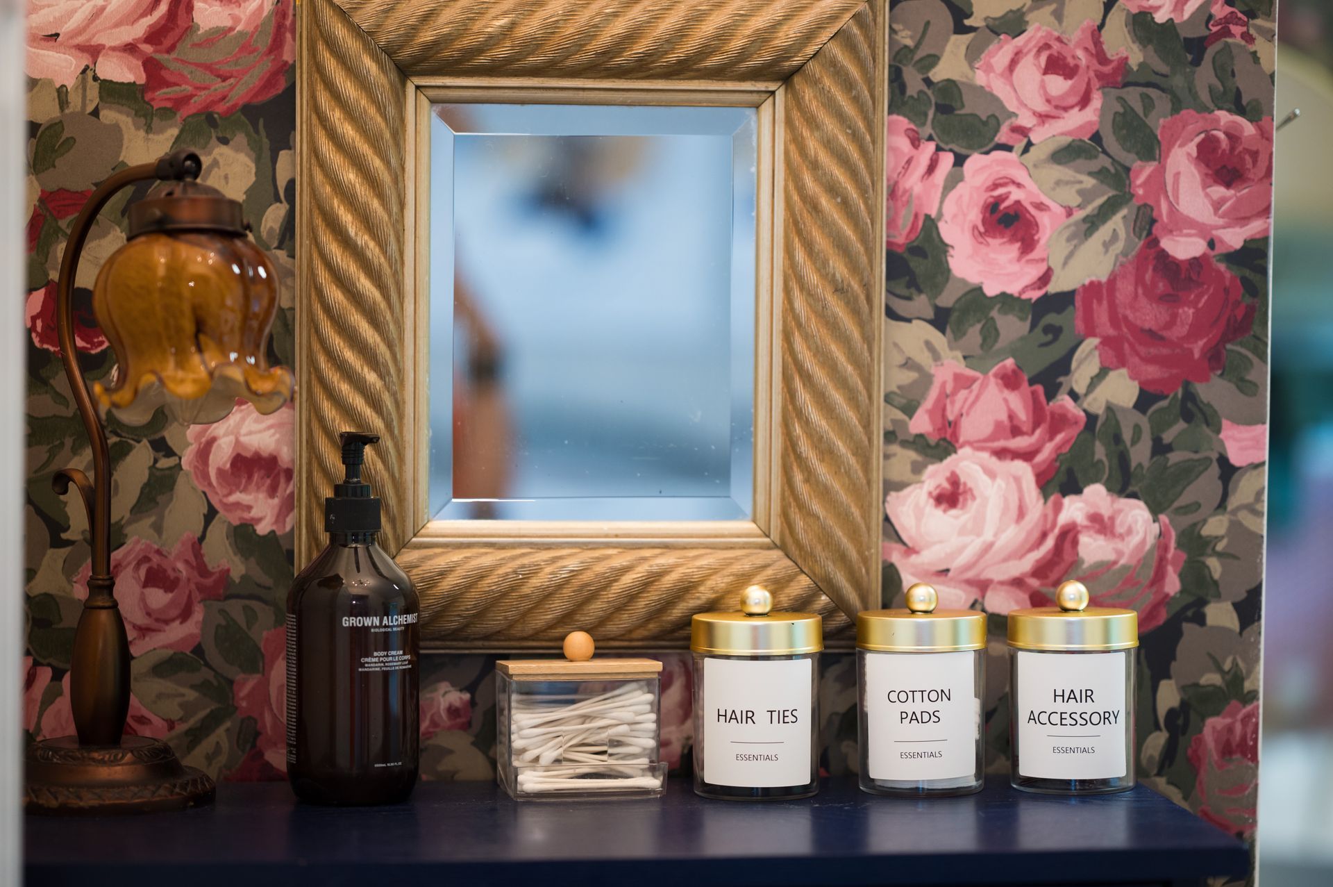 Mirror with floral wallpaper background, lamp, and jars on a blue shelf.