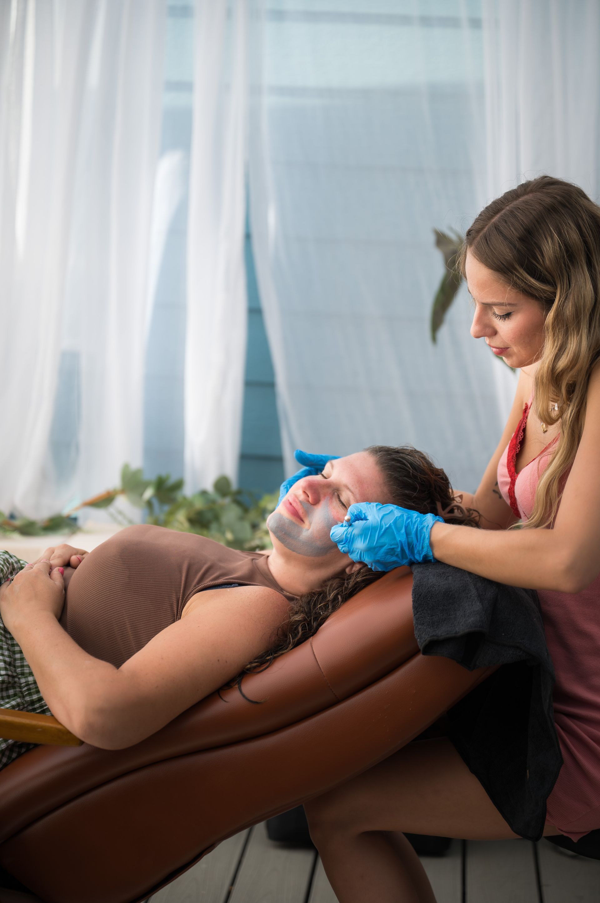 Person receiving a facial treatment; esthetician applying mask, both in spa setting with blue gloves.