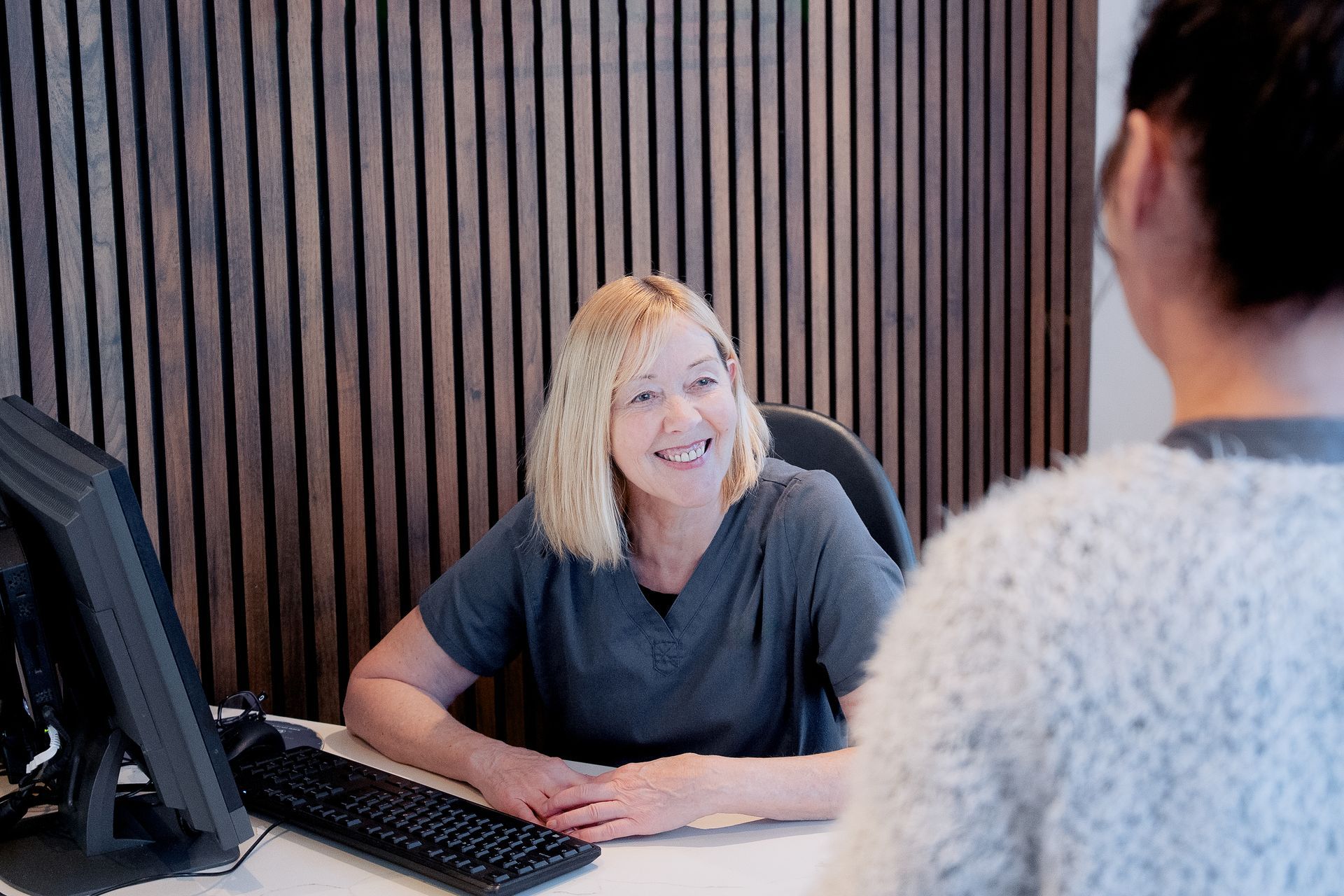 Woman at a desk smiles at another person. Wooden paneling. Grey scrubs. Keyboard and computer visible.