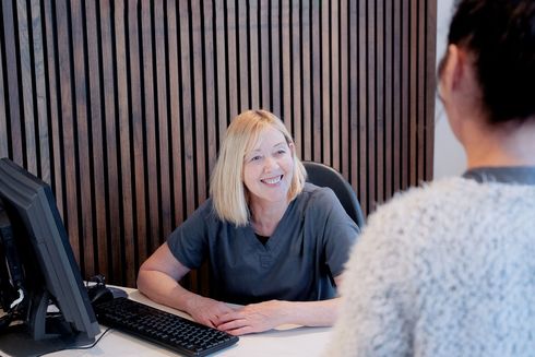 Receptionist smiling, facing patient at a desk, wood-paneled wall backdrop.