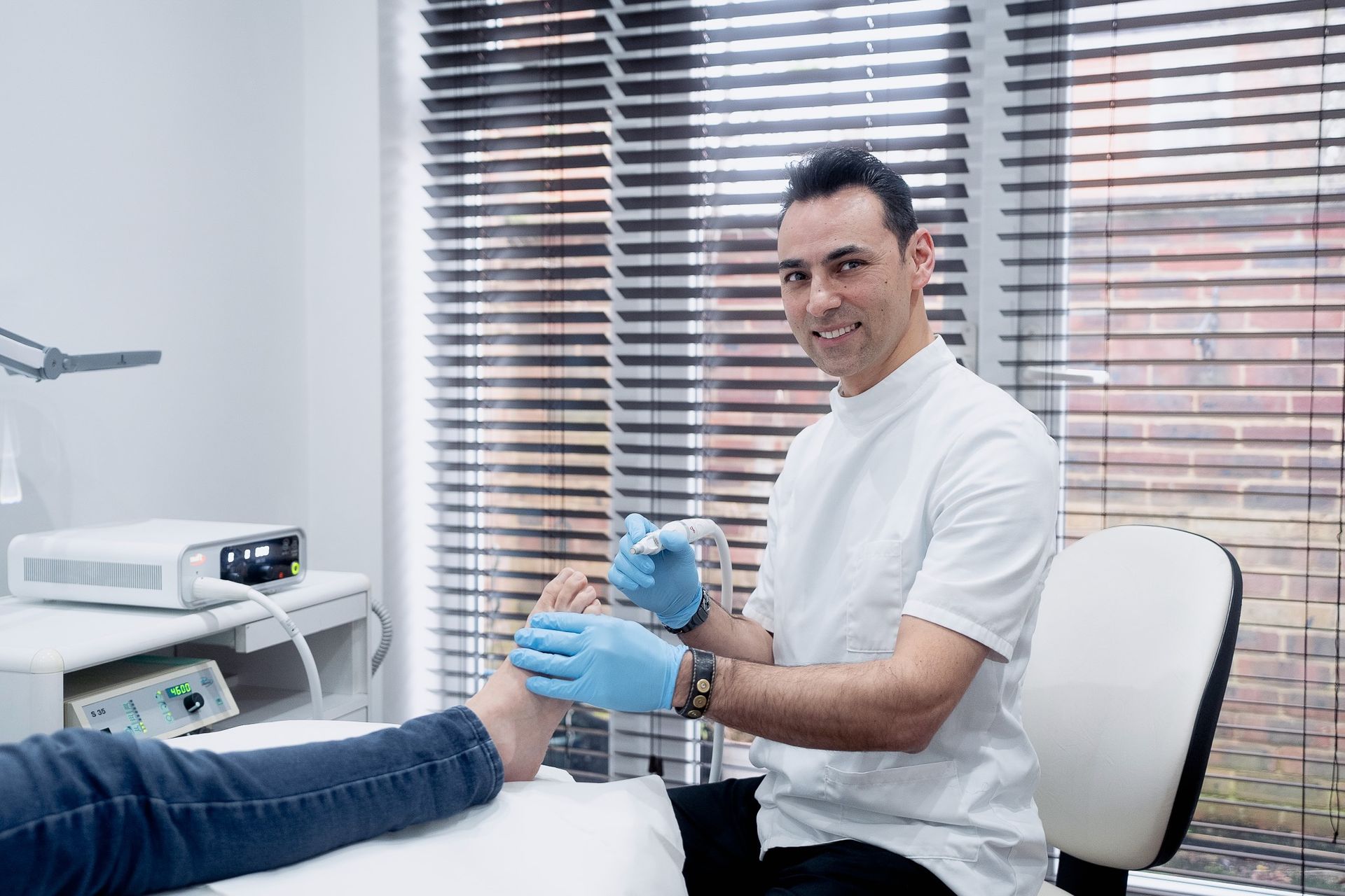 Podiatrist examining a patient's foot in an office; window with blinds in the background.