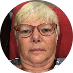 Person with short blonde hair and glasses, sitting in front of a red background.