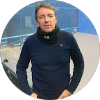 Man in blue shirt and neck gaiter stands in front of a squash court.