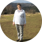 Woman standing in a field wearing a striped shirt and pants, with a forest in the background.
