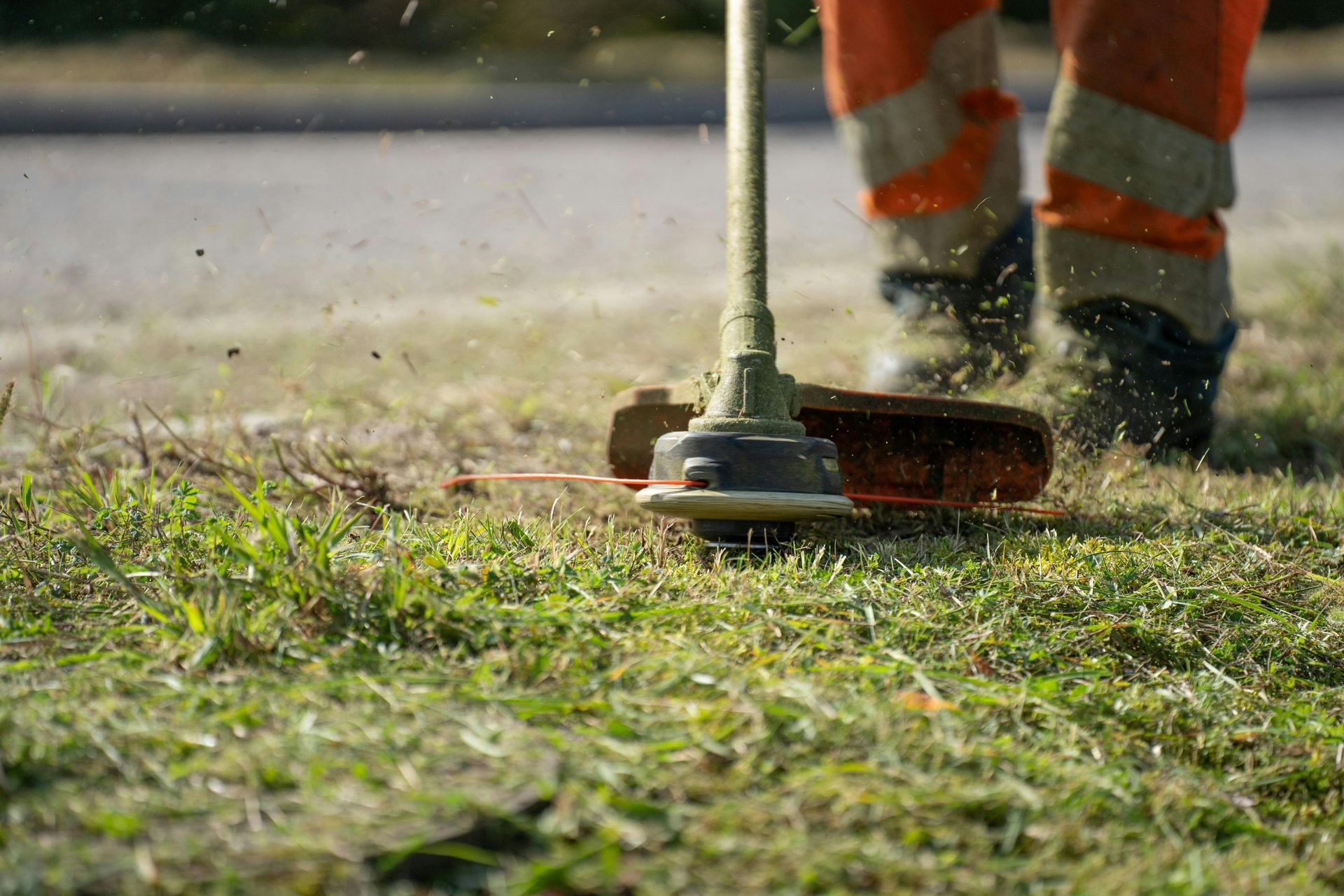 Weed trimmer cutting grass near a curb, with worker’s orange reflective pants visible in the background