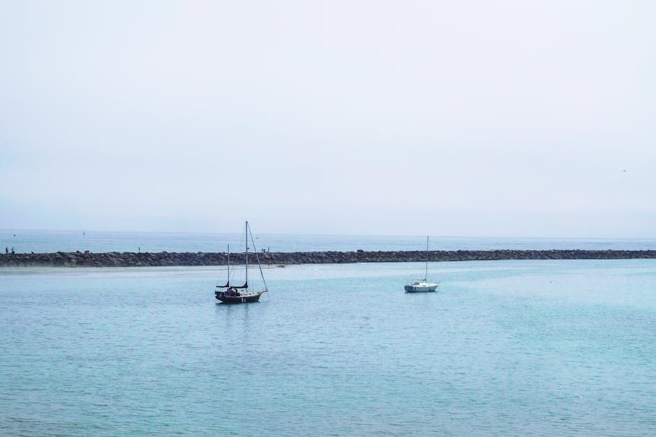 Two sailboats in a calm, turquoise harbor on an overcast day, with a rock wall in the distance.