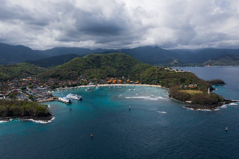 Aerial view of a harbor town on a bay, with boats, turquoise water, and green hills under a cloudy sky.