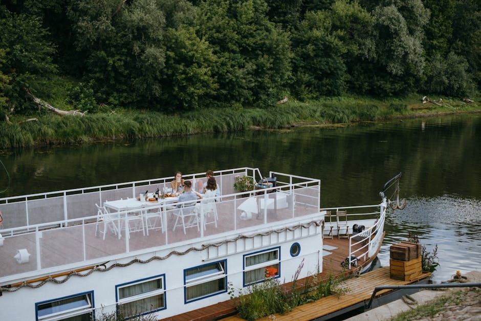White houseboat with people dining on the deck, docked on a river, surrounded by trees.