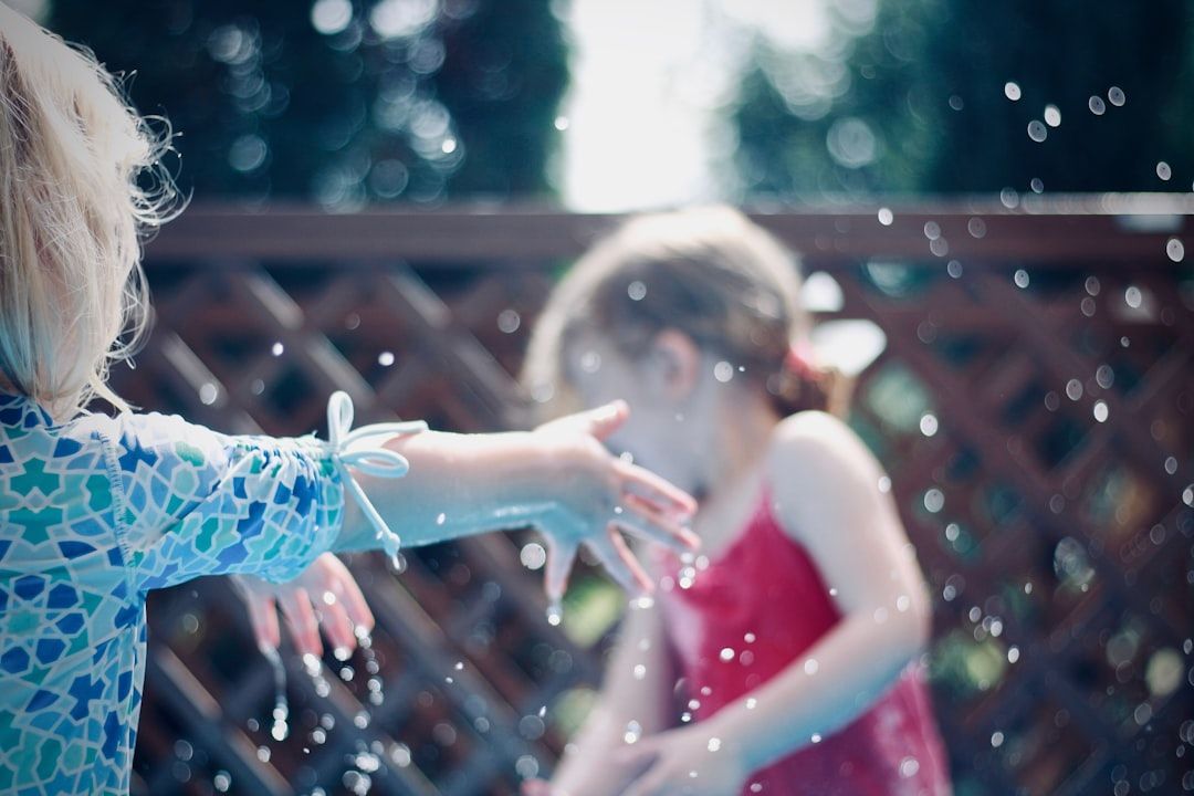 Two young girls playing in water, one in a blue shirt, the other in a red swimsuit. Water droplets in the air.