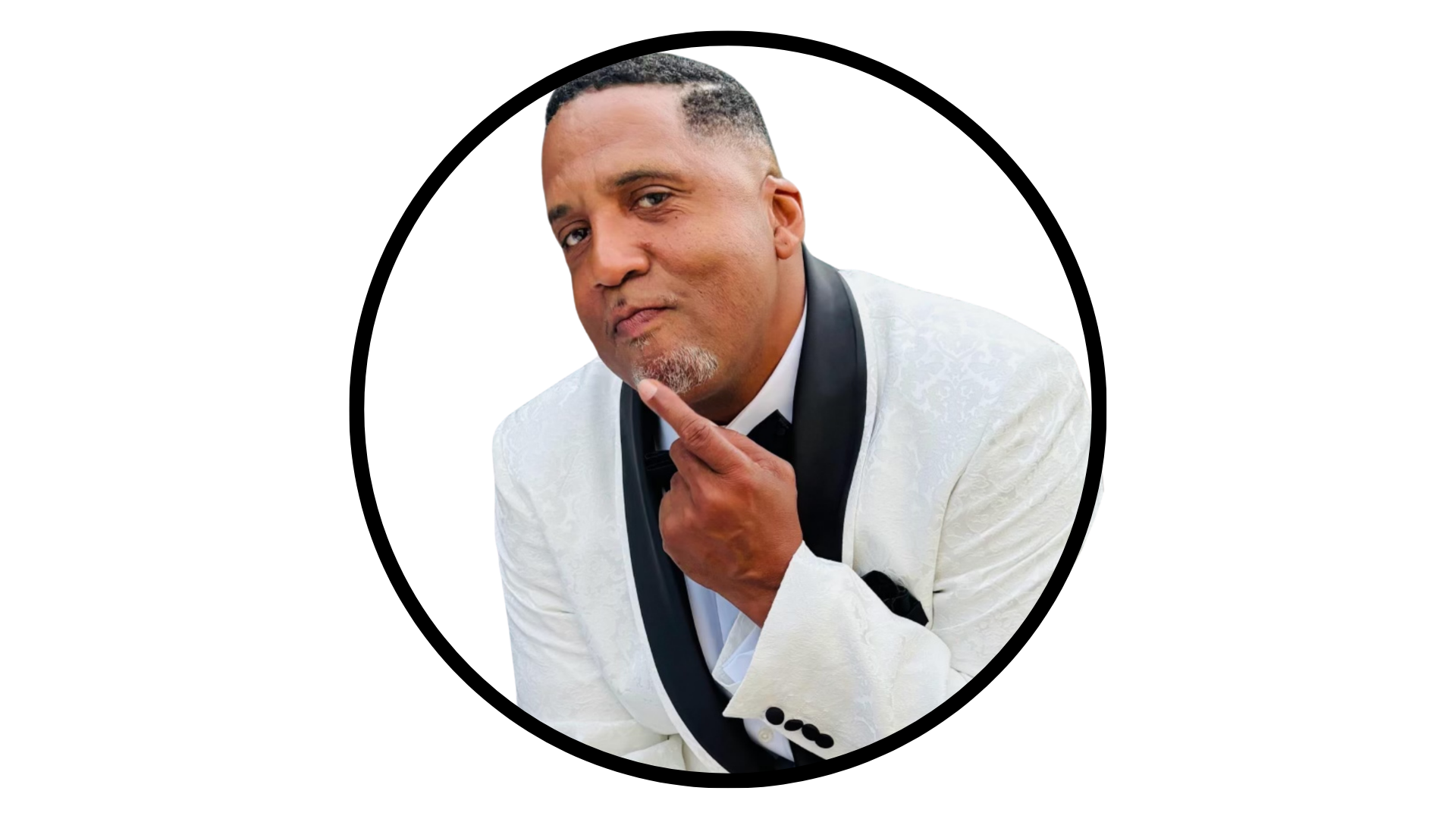 A man in a white tuxedo jacket with a black lapel, looking directly at the camera with his index finger near his chin.