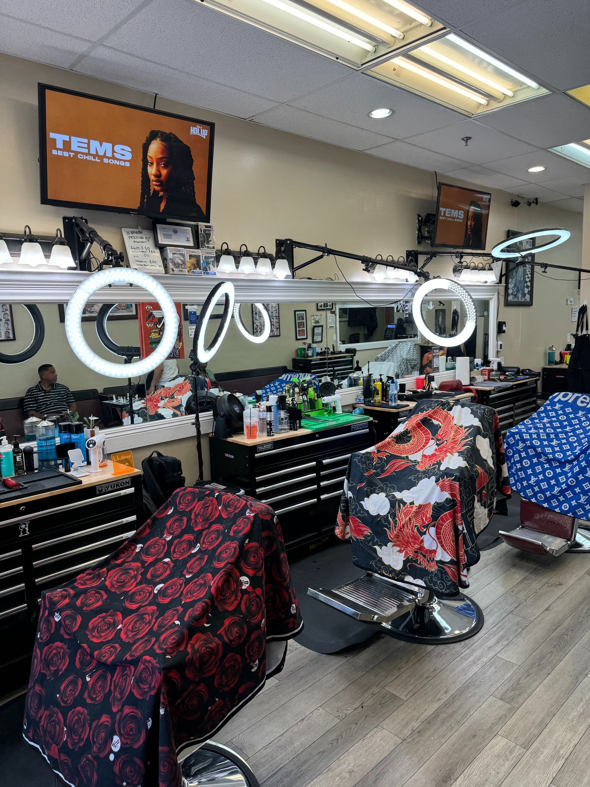 A barber shop interior featuring three chairs covered in patterned capes, each equipped with a professional ring light.
