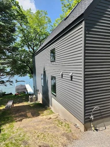 Gray-sided house on a waterfront property; two windows, dock, trees, blue water, and bright sky.