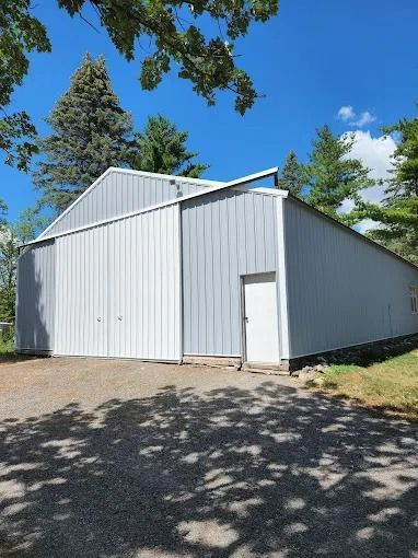 Gray metal barn with sliding doors and a white door on a gravel lot, under a blue sky.