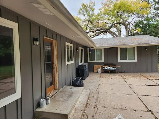 Gray house exterior with white trim, concrete patio, and large tree in the background.