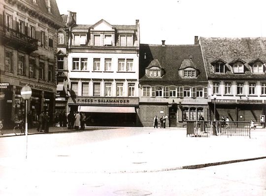 Gebäude auf einem Platz. Ein Schild trägt die Aufschrift „KIEFER – SILBERWAREN“. Personen und ein Straßenschild