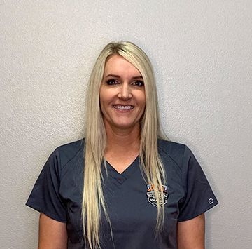 A woman with long blonde hair is wearing a scrub top and smiling.