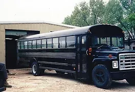 a black school bus is parked in a dirt lot in front of a garage .
