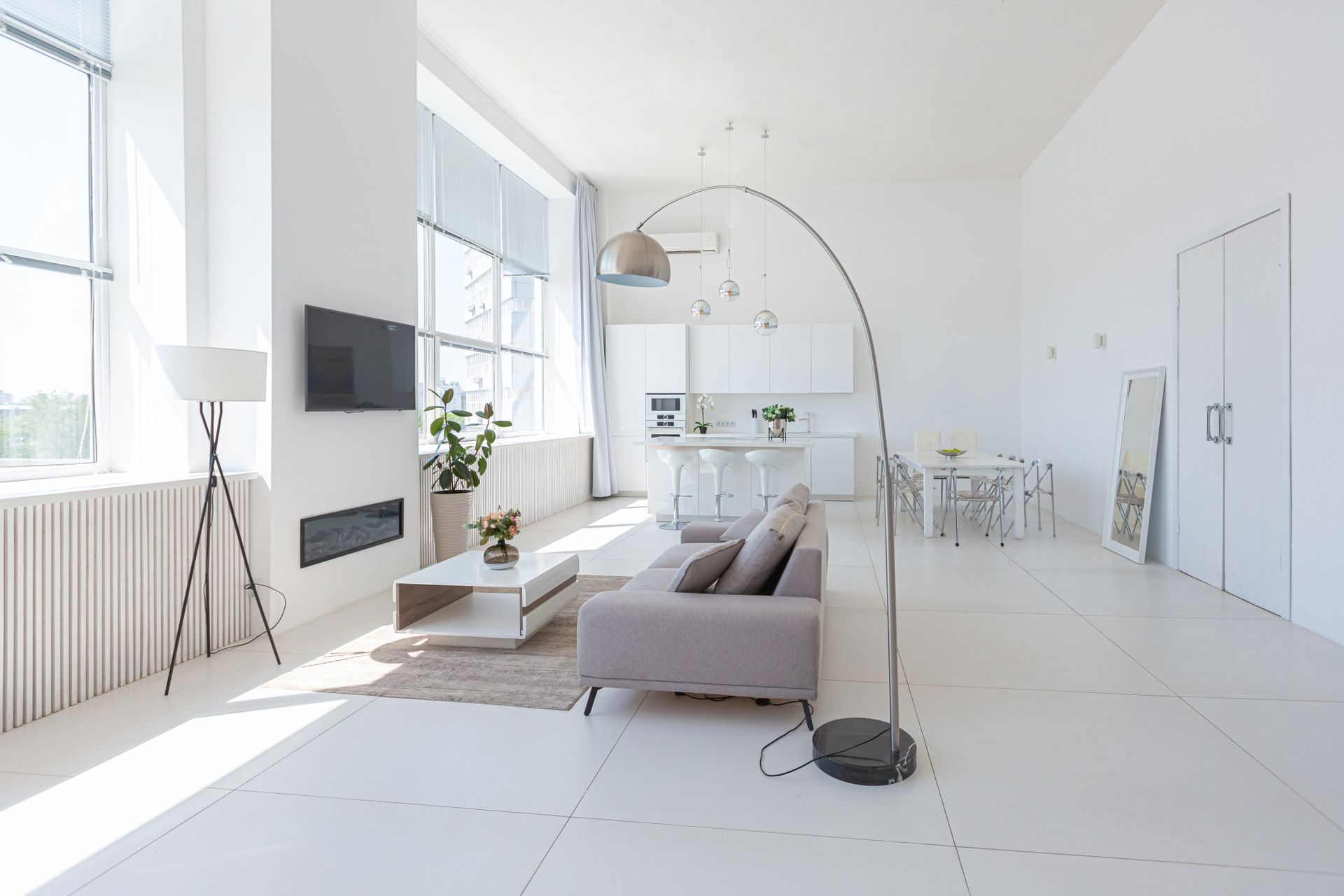 A bright, modern open-concept living area with white walls, white tile flooring, a gray sofa, and a tall arc floor lamp.