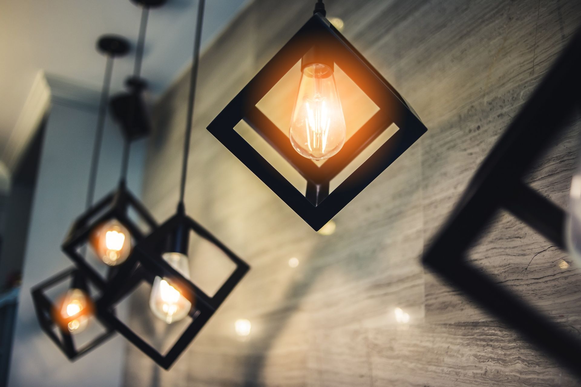A series of geometric, black-framed pendant lights with glowing Edison bulbs hanging against a textured gray wall.