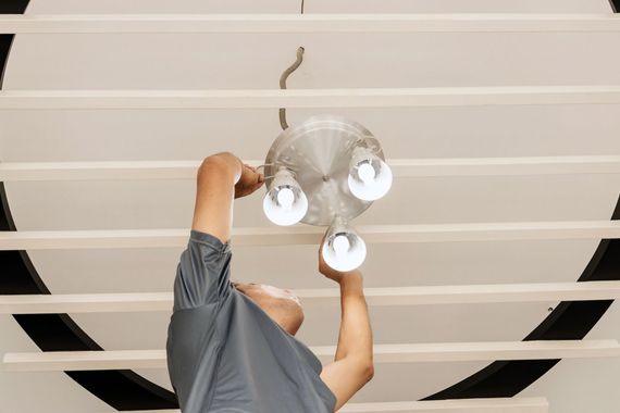 A person in a gray shirt reaching up to install or adjust a modern three-bulb ceiling light fixture.
