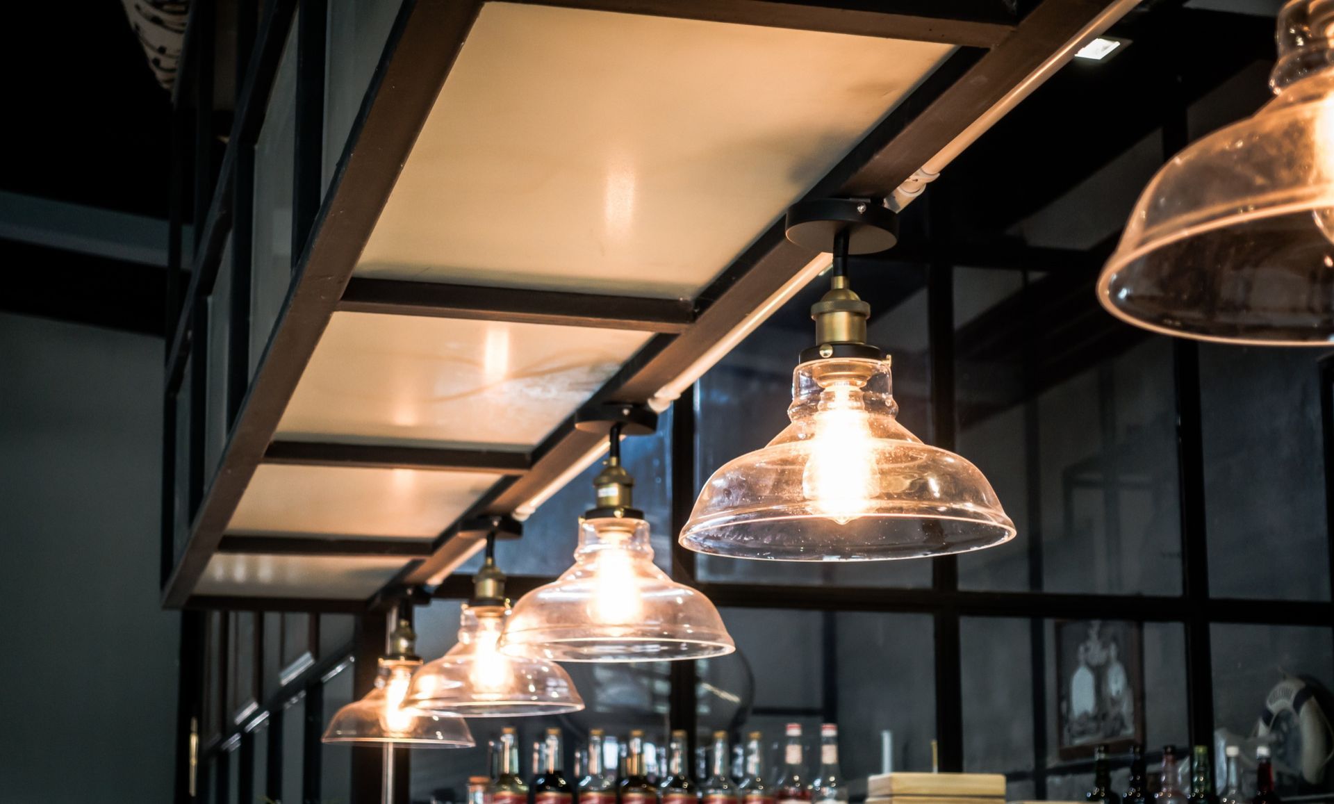 A row of vintage glass pendant lights with warm glowing bulbs hanging from a dark metal ceiling rack in a bar setting.