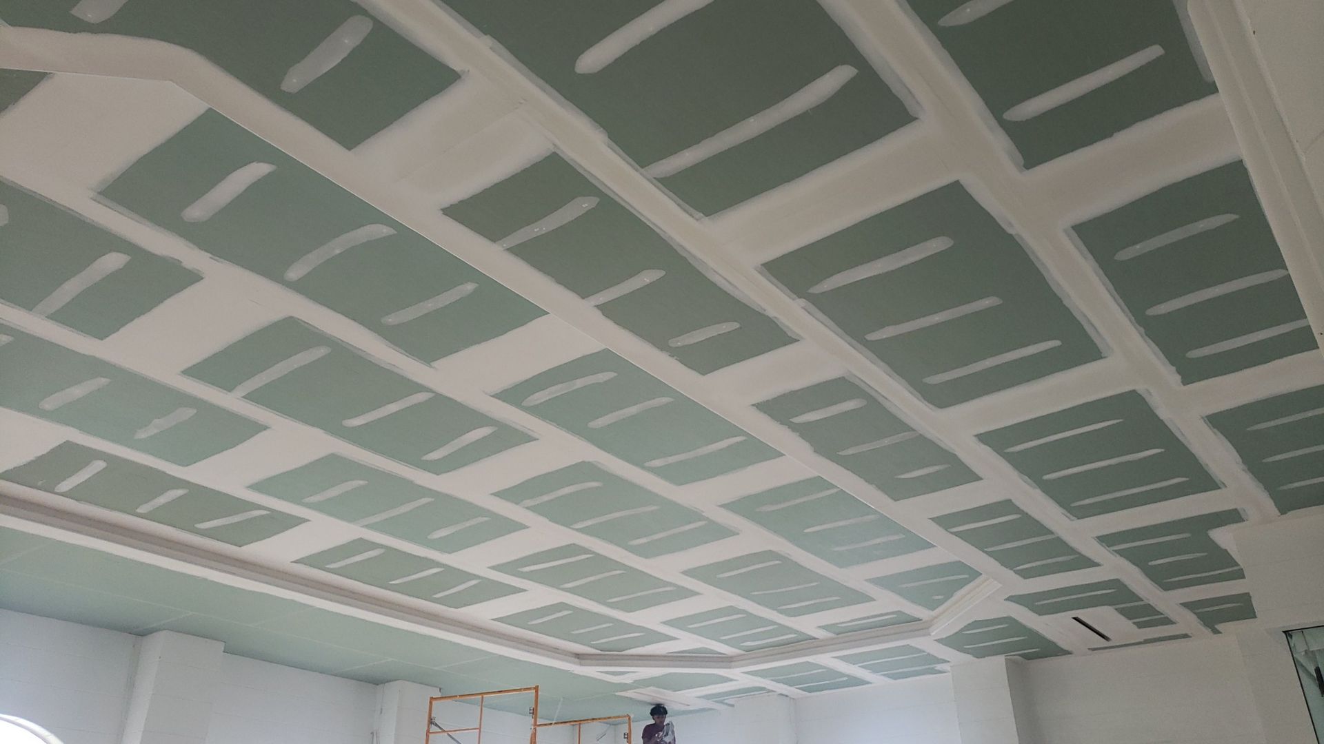 A ceiling under construction, featuring green drywall panels secured with white joint compound in a grid pattern.