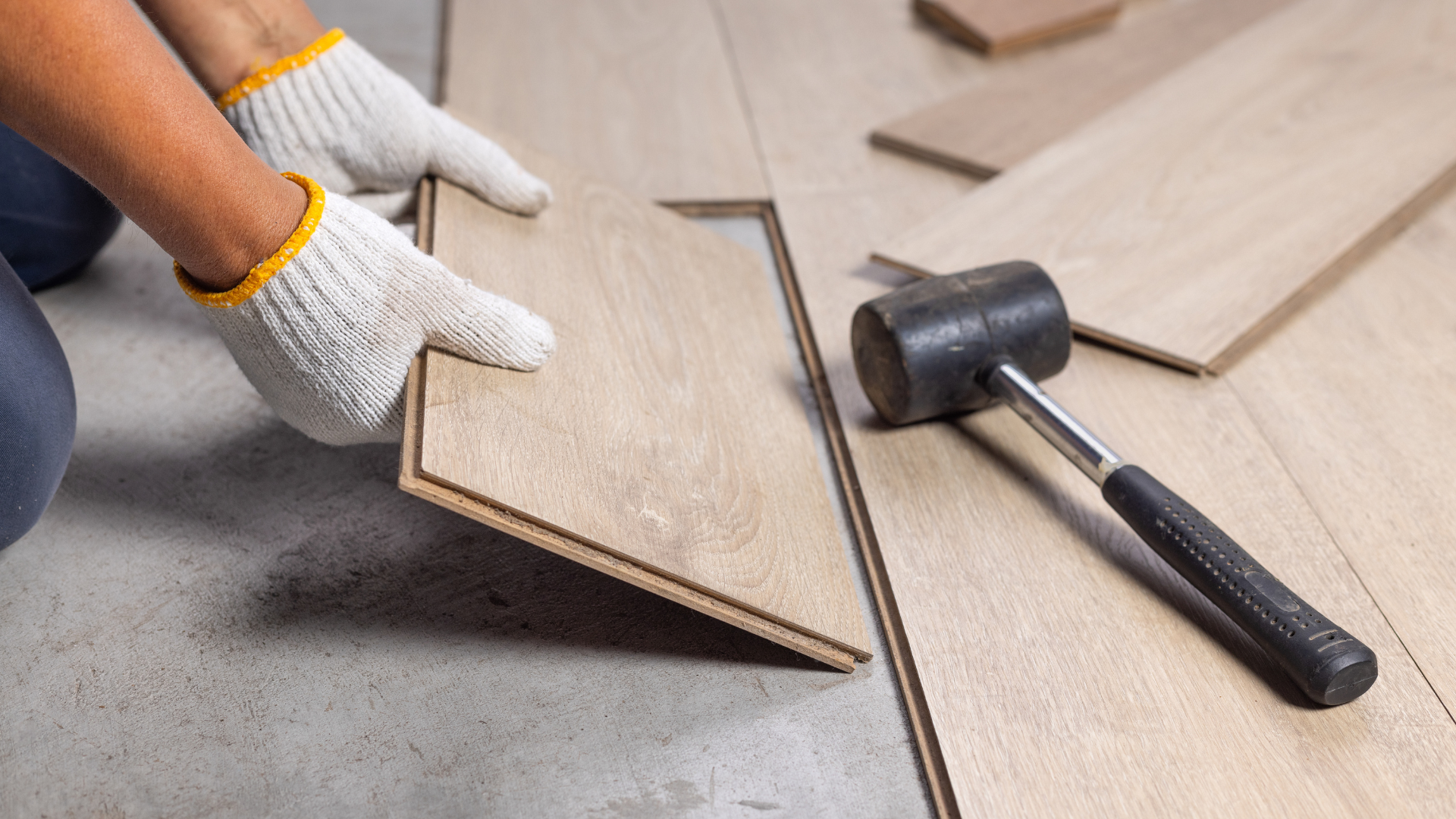Worker wearing gloves installs a light-wood laminate flooring plank with a rubber mallet nearby.