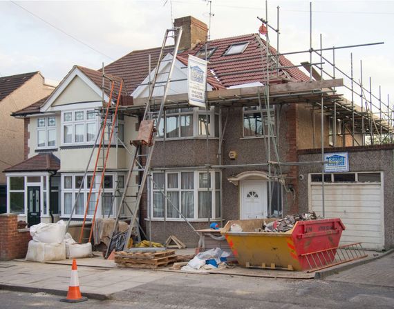 A semi-detached house under renovation with scaffolding covering the roof and front, and a construction skip out front.