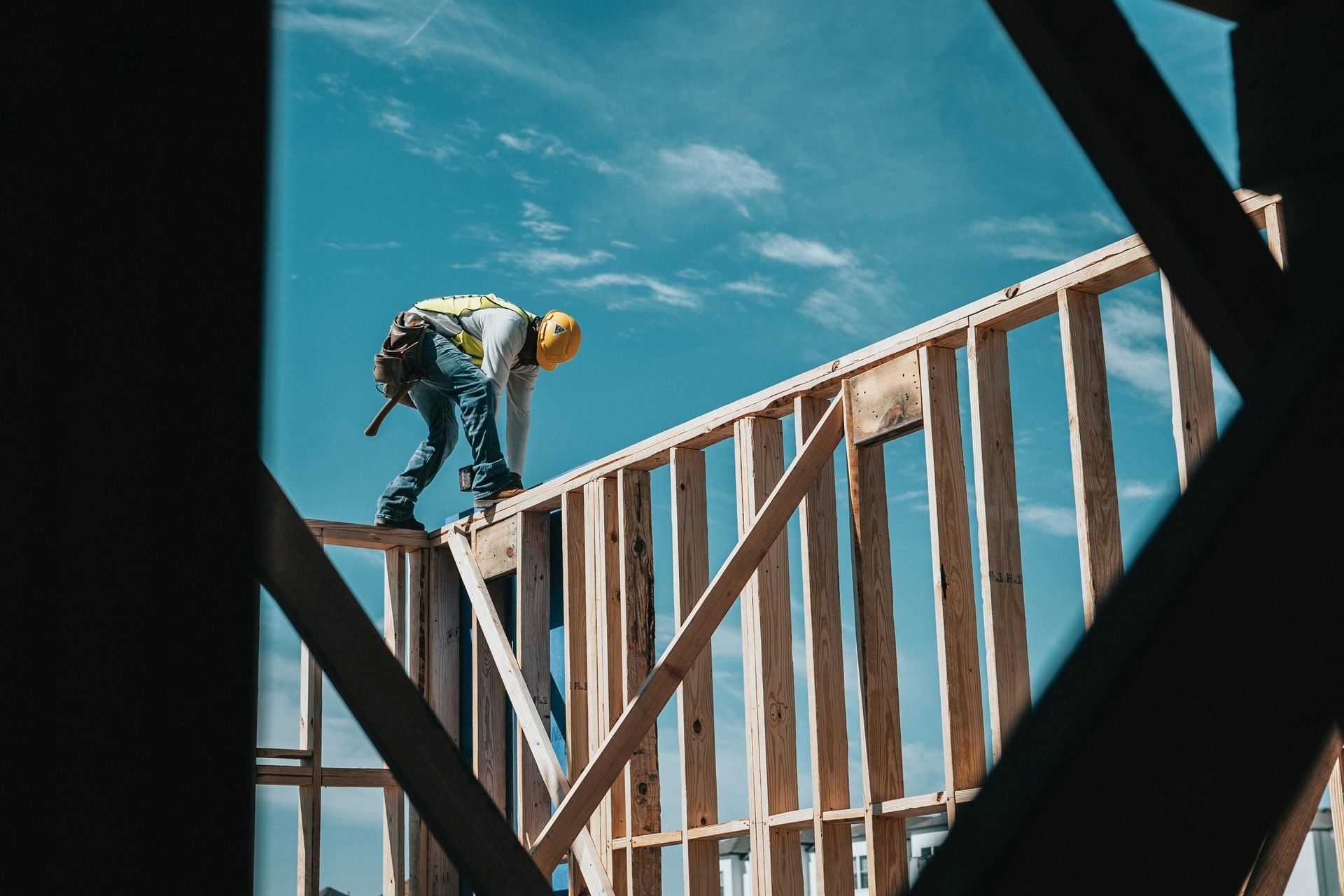 A construction worker in a yellow hard hat balances on the wooden frame of a building under a bright blue sky.