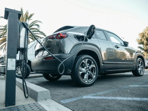 A dark gray electric car parked in an outdoor lot, charging via a cable connected to a station on the left.