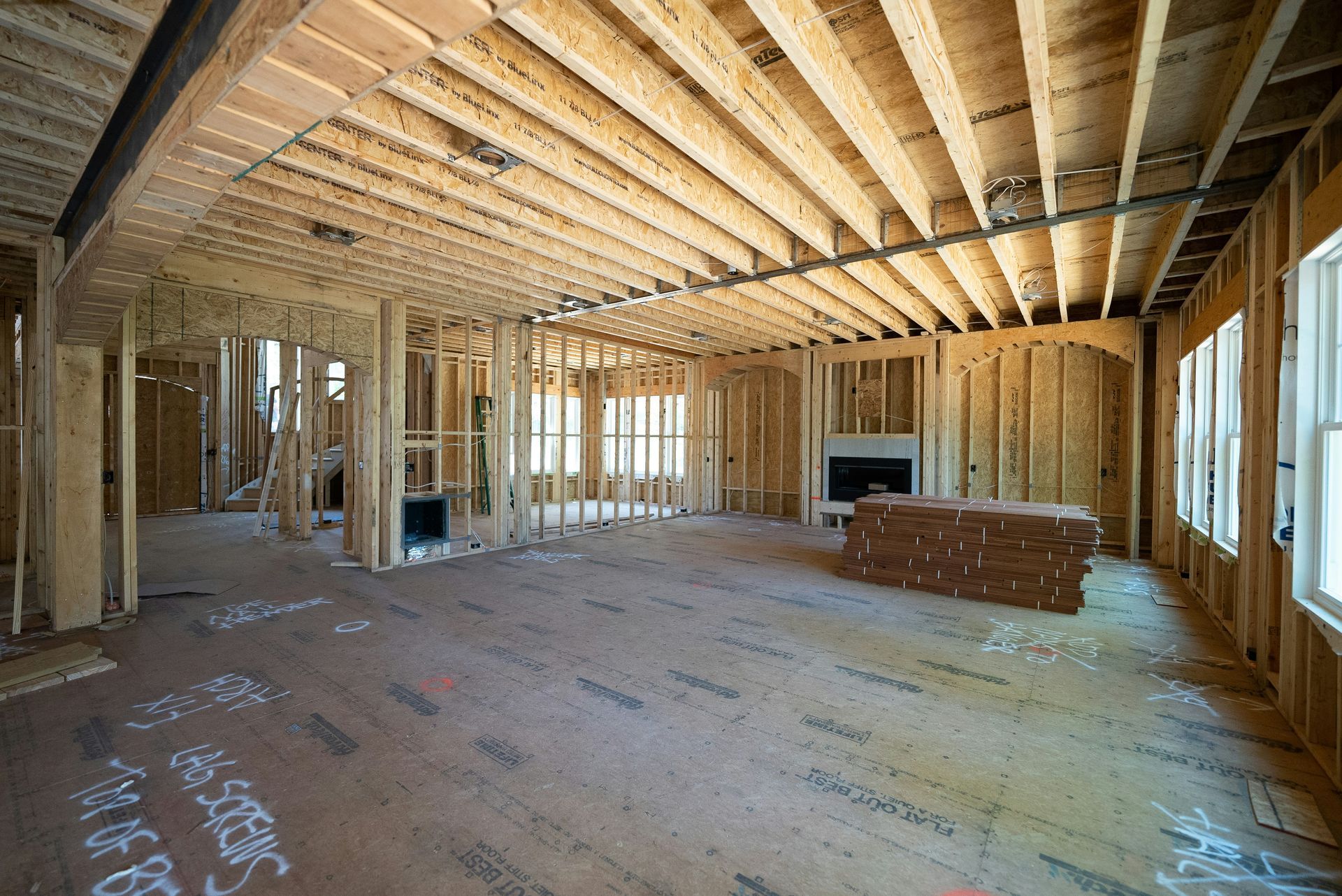 An open-concept house under construction, showing wooden wall studs, exposed ceiling joists, and a fireplace frame.