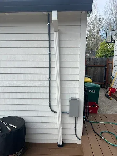 Electrical conduit runs along the exterior wall of a white house, connecting to a grey junction box above a wooden deck.