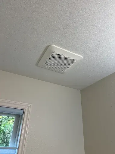 A white square ventilation fan grille mounted on a white ceiling next to a window.