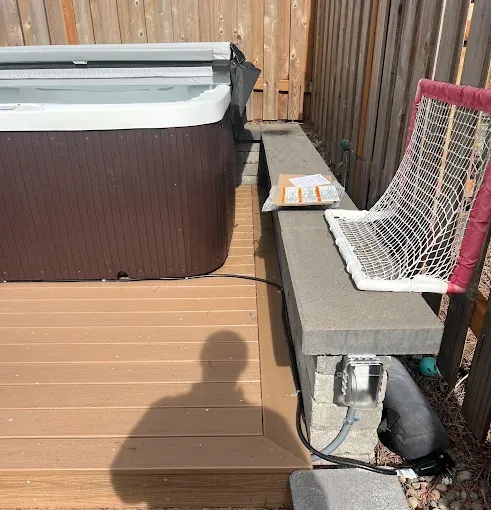 A brown hot tub sits on a wooden deck next to a concrete block ledge with an electrical outlet and a small hockey net.