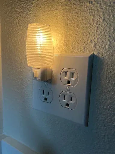 A glowing, ridged white nightlight is plugged into the top-left outlet of a four-outlet wall plate on a textured wall.