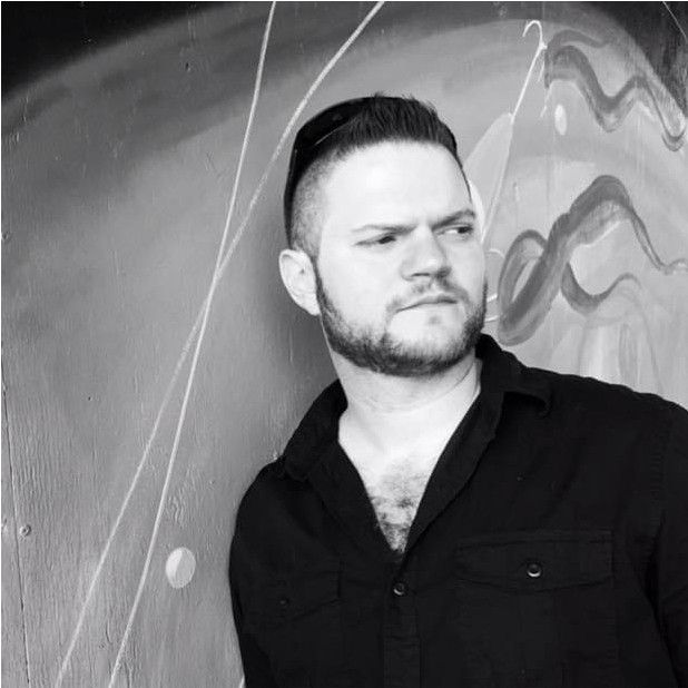 Man with dark hair and beard, wearing a black shirt, looking to the side, against a painted wall.