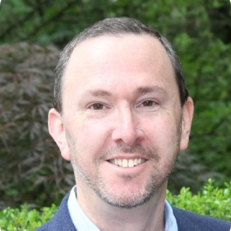Man in blue blazer smiles in front of green foliage.